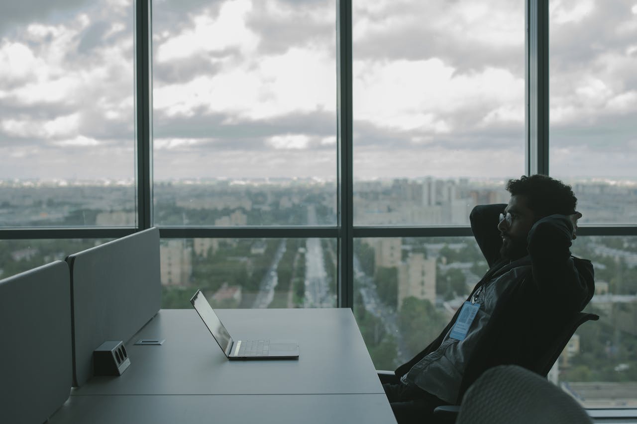 services-03 A man sits in an office with hands on head in front of a laptop, overlooking a cityscape.