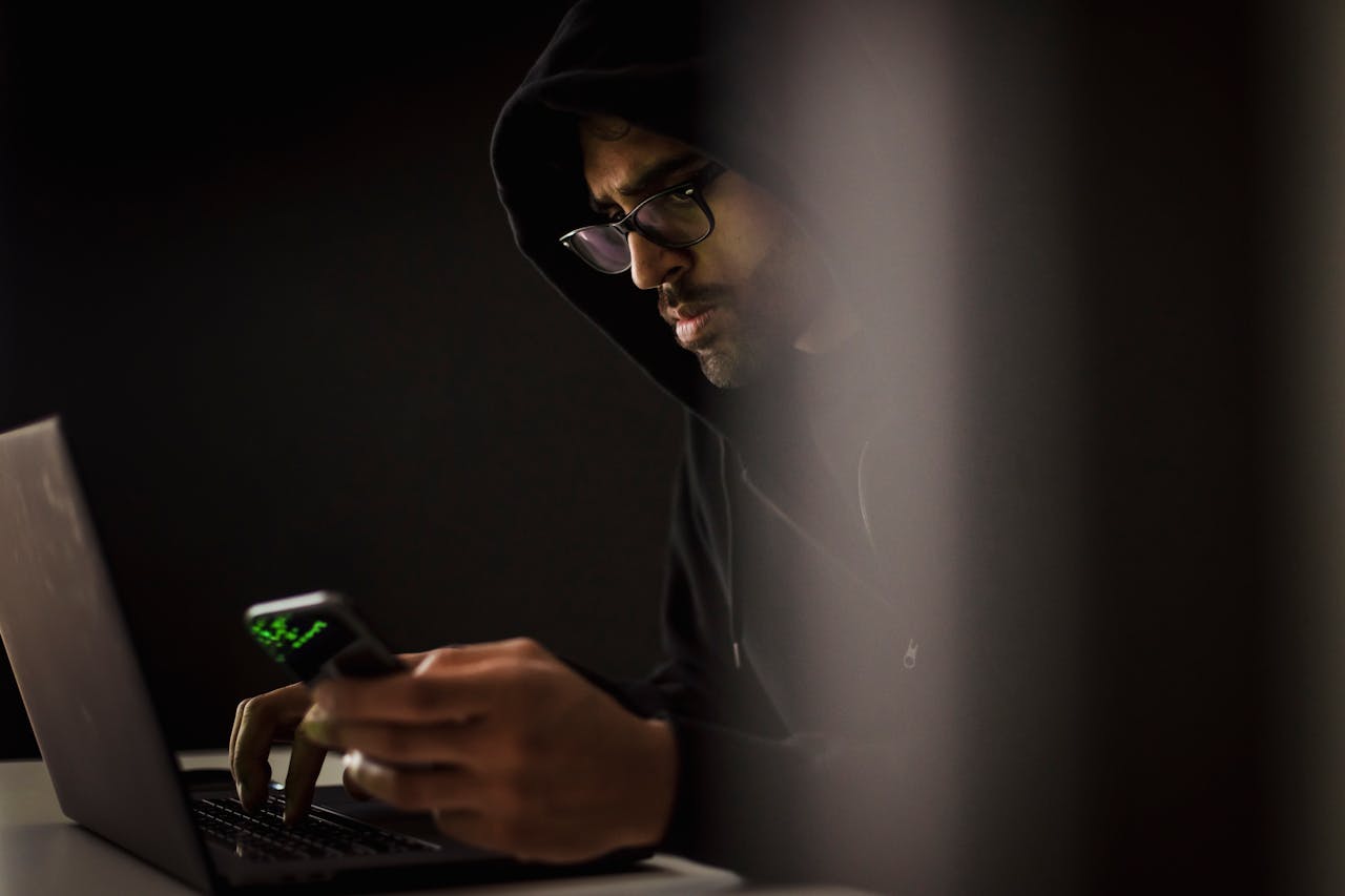 services-02 Man in hoodie using laptop and smartphone in dark room, focused and concentrated.